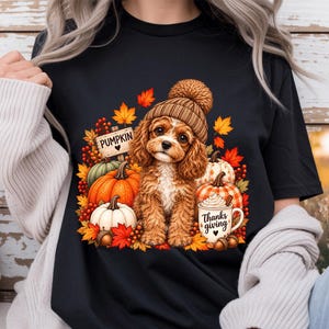 May include: A black t-shirt featuring a graphic of a brown puppy wearing a tan knit pom-pom hat. The puppy is surrounded by colourful autumn leaves, pumpkins, and a mug with the text "Thanks giving" and a sign that says "PUMPKIN".