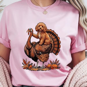 May include: A light pink t-shirt featuring a graphic illustration of a brown curly-haired dog sitting on the back of a large turkey. The design includes autumn-themed elements like orange leaves and small pumpkins at the base of the turkey.