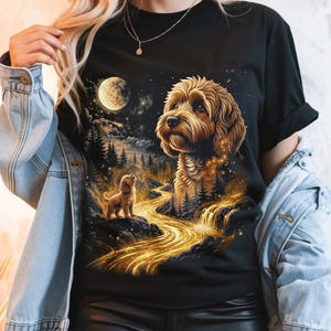 Puede incluir: Una camiseta negra de manga corta con un estampado gráfico de un perro de color dorado mirando hacia una luna llena en un cielo estrellado. El diseño incluye un río dorado brillante que fluye a través de un paisaje forestal oscuro.