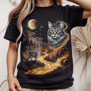 May include: A black short-sleeved t-shirt featuring a graphic print of a tabby cat face and a full-body tabby cat standing on a rock. The scene includes a glowing golden river, a dark forest, a waterfall, and a large moon in a starry night sky.