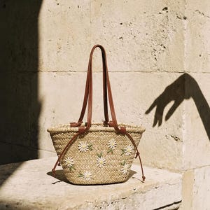 May include: A woven straw basket bag featuring brown leather handles and embroidered white daisy flowers with green stems. The bag has a tapered shape with a wide opening and thin leather straps extending down the sides of the woven body.