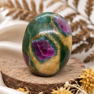 May include: A polished, egg-shaped stone featuring a vibrant mix of deep magenta, forest green, and golden-yellow mineral inclusions. The surface is smooth and reflective, showcasing distinct colour zones and natural patterns throughout the stone.