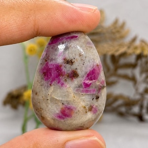 Pode incluir: Uma pedra polida em forma de lágrima com uma base lisa cinza-claro e inclusões irregulares proeminentes de depósitos minerais em magenta vibrante e rosa escuro espalhados por toda a superfície da gema.
