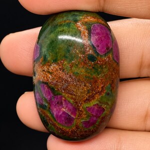 Puede incluir: Una piedra preciosa pulida de forma ovalada con una superficie moteada en tonos verde oscuro y marrón tierra, acentuada por inclusiones vibrantes e irregulares de color magenta distribuidas por toda la superficie lisa, brillante y redondeada.