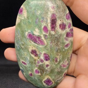 Peut inclure: Une pierre polie de forme ovale présentant une surface lisse et marbrée vert clair, incrustée de nombreuses inclusions cristallines distinctes et irrégulières de couleur magenta profond et rouge rubis sur toute la surface.