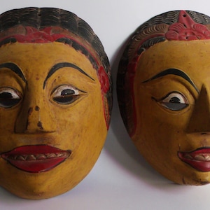 Peut inclure: Deux masques en bois avec des visages peints. Les masques ont les cheveux noirs, les lèvres rouges et la peau jaune. Les masques sont décorés de peinture rouge et brune.