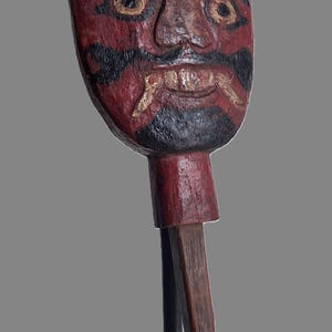 Può includere: Una maschera in legno intagliato a mano con base rossa, caratterizzata da sopracciglia, baffi e barba dipinti in nero. La maschera presenta un bordo superiore a corona, occhi prominenti e denti visibili su una base nera.