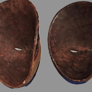 Puede incluir: Dos máscaras de madera de forma ovalada mostradas desde el lado interior. Las máscaras presentan un acabado marrón oscuro, liso y pulido con veta de madera visible. Cada máscara tiene una ranura ocular horizontal en el centro.