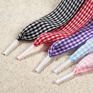 Op de afbeelding: Een set van vijf platte veters met een vichy-ruitpatroon in zwart, rood, paars, lichtblauw en lichtroze. Elke veter is afgewerkt met een gladde, witte plastic nestel aan het uiteinde, wat zorgt voor een nette en functionele afwerking.