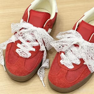 Op de afbeelding: Een paar rode suède sneakers met witte kanten veters die in grote strikken zijn gestrikt. De sneakers hebben een laag model, een gladde rode tong, een crèmekleurige voering aan de binnenkant en een platte bruine rubberen zool.