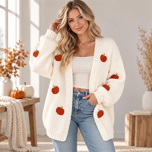 May include: An off-white, chunky knit open-front cardigan featuring a repeating pattern of small, embroidered orange pumpkins with green stems scattered across the front panels and sleeves, finished with ribbed cuffs and a ribbed hemline.