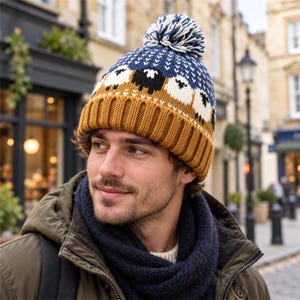 May include: A knit beanie featuring a mustard yellow ribbed cuff and a blue crown decorated with a repeating pattern of white and black sheep. The hat is topped with a blue and white pom-pom and includes a small white geometric design.
