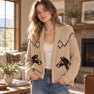 May include: A beige knit cardigan featuring a shawl collar, long sleeves, and a dark brown horse head graphic on each side. The sweater includes a black zigzag pattern across the chest and shoulders, with a ribbed hem and cuffs.