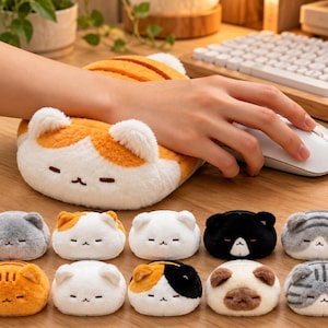 May include: A plush cat-shaped wrist rest with soft white and orange fur, featuring embroidered closed eyes and a small mouth. The collection includes ten variations of these small, rounded, fuzzy cat-themed wrist supports in colors like gray, black, white, and tabby patterns.