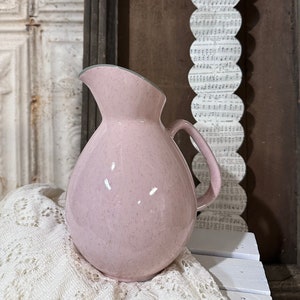 May include: A pink speckled ceramic pitcher with a gray rim and a handle. The pitcher is sitting on a white lace tablecloth.