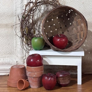 May include: A white wooden stool with a woven basket, a green apple, and four red apples. The basket is sitting on the stool and the apples are arranged around the base of the stool.