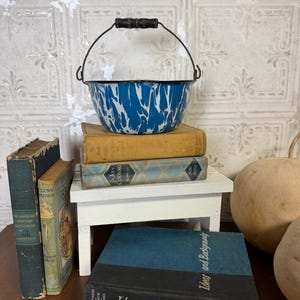May include: A blue and white enamelware bowl with a black handle sits atop a stack of vintage books. The bowl has a black rim and a metal handle with a wooden grip. The books are various colors and sizes, adding to the rustic aesthetic.