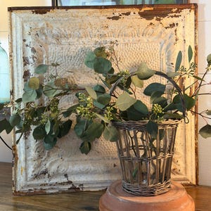 May include: A rustic display featuring a woven basket vase holding eucalyptus branches, set on a terracotta base. The arrangement is placed in front of a distressed, white, embossed metal panel and a framed painting.
