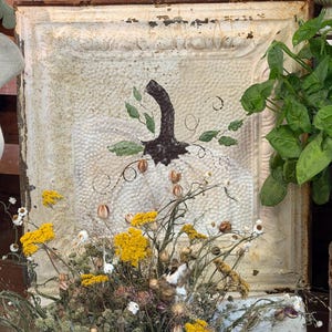 May include: A square, off-white, distressed metal ceiling tile featuring a painted dark brown pumpkin silhouette with green leaves and swirling vines. The tile has a textured, embossed border and surface, showing signs of rust and age.