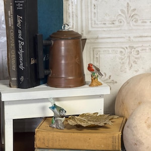 May include: A vintage copper coffee pot with a glass knob sits on a white shelf next to a stack of old books. A small ceramic bird figurine and a gold leaf-shaped dish rest on top of the books. The books include titles like "Ideas and Backgrounds" and "Joy of Cooking".
