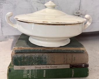 May include: A white ceramic serving dish with a gold rim and lid. The dish is sitting on a stack of three green books. The top book has the title "Snug in the Snow" printed on the spine.