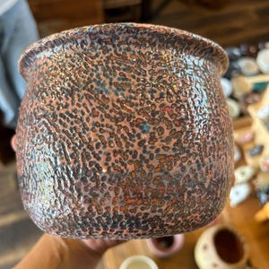 以下が含まれることがあります： 銅色、茶色、濃い青色の斑点模様の釉薬が施された、質感のあるくぼみのある表面を持つ丸みを帯びた陶器の鉢。この容器は、幅広でわずかに広がった縁と、深みのある球根状の本体を備えており、全体的に不均一な仕上がりになっています。