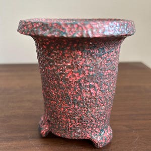 以下が含まれることがあります： 鮮やかなピンクとダークチャコールグレーのまだら模様が特徴的な、クレーターのような質感の表面を持つ小さな広がったセラミック製の鉢。この容器は3本の短い頑丈な脚で支えられており、幅広でわずかに張り出した縁があります。