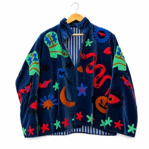Peut inclure: Une veste en velours bleu marine ornée de motifs brodés colorés, notamment des bottes de cow-boy vertes, des serpents rouges, des croissants de lune orange, des fleurs bleues et des yeux. La doublure intérieure présente des rayures verticales.