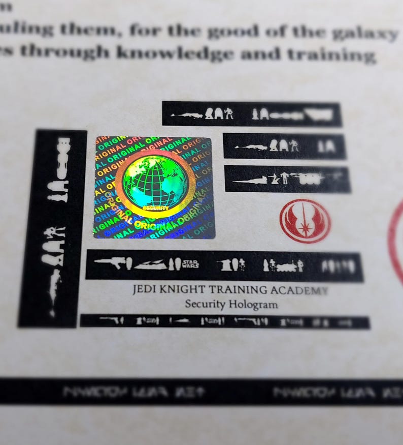 May include: A holographic sticker with a globe design and the text "ORIGINAL" repeated around the globe. The sticker also has the text "JEDI KNIGHT TRAINING ACADEMY" and "Security Hologram" below it.