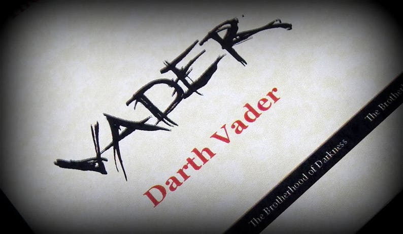 May include: A black and white illustration of the word "Vader" with a red and white text overlay that reads "Darth Vader" and "The Brotherhood of Darkness" in the bottom right corner.