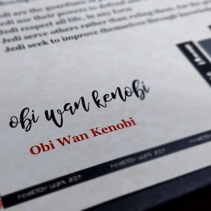 May include: A white piece of paper with black text that reads "The following are the guidelines to define... I. Jedi use their powers... II. Jedi respect all life... III. Jedi serve others rather than ruling through knowledge... IV. Jedi seek to improve themselves..." and a black and white image of a Jedi Knight. The text "obi wan kenobi" is written in black cursive and "Obi Wan Kenobi" is written in red below it.