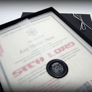 May include: A black framed certificate with a white background and red text that reads "The Brotherhood of Darkness Recognizes Any Name Here as a Sith Lord". The certificate has a wax seal with a Star Wars Stormtrooper helmet design.