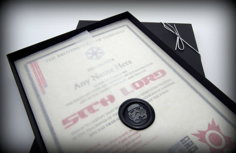 May include: A black framed certificate with a white background and red text that reads "The Brotherhood of Darkness Recognizes Any Name Here as a Sith Lord". The certificate has a wax seal with a Star Wars Stormtrooper helmet design.