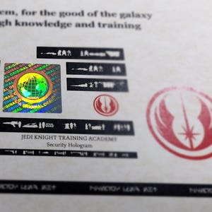 May include: A close-up of a holographic sticker with the text "JEDI KNIGHT TRAINING ACADEMY" and the words "Security Hologram". The sticker features a colorful globe with the words "ORIGINAL" repeated around it. The sticker is on a white background with a red and white Jedi symbol.