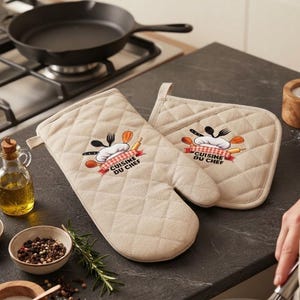 Peut inclure: Un gant de cuisine matelassé beige et une manique carrée assortie, tous deux ornés d'un motif de toque de chef, d'ustensiles de cuisine et du texte "CUISINE DU CHEF" en rouge et noir. Les articles présentent un motif piqué en losanges.