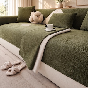 May include: A sofa covered in a textured olive green fabric with a waffle-knit pattern. The sofa features matching olive green cushions, a cream-colored decorative knot pillow, and a white mug with a butterfly accent. A pair of cream slippers sits nearby.