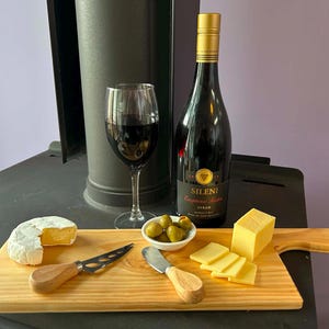 Könnte beinhalten: Ein hölzernes Servierbrett mit einem runden Brie, einem Block gelbem Cheddar, Cheddar-Scheiben, einer kleinen Schale mit grünen Oliven und zwei Käsemessern mit Holzgriffen. Dahinter stehen ein Glas Rotwein und eine Flasche Sileni-Wein.