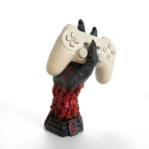 May include: A cream-colored video game controller held by a dark, textured hand sculpture. The base of the sculpture features a cluster of red, sculpted faces and skulls, with a red symbol on the palm and a matching red symbol on the front base.