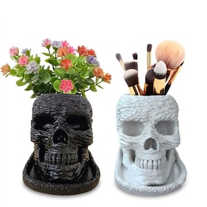 May include: A black skull-shaped planter containing small pink, orange, and purple artificial flowers, and a white skull-shaped holder containing several makeup brushes with brown and tan bristles, both resting on matching circular bases.