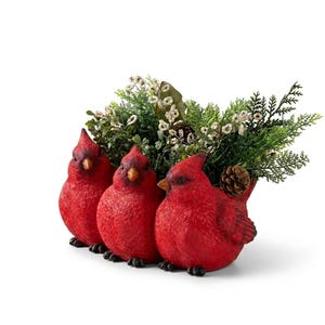 May include: A decorative figurine featuring three bright red cardinal birds positioned side-by-side. The birds have orange beaks and black feet. Behind the birds is an arrangement of green foliage, small white flowers, and a single brown pinecone.