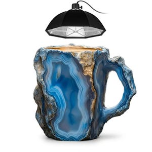 May include: A mug with a textured exterior resembling blue and white agate stone with gold-colored accents. The mug features a rugged, natural-looking surface and a handle. Above the mug, a black photography softbox light is suspended.