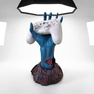 May include: A blue, textured hand sculpture emerging from a dark, rocky base with a red symbol, holding a white video game controller. The controller features two analog sticks, a directional pad, and buttons labeled with the letters A, B, X, and Y.