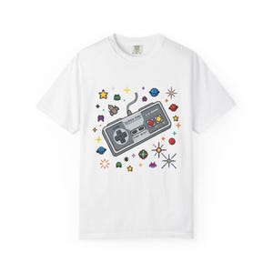 May include: A white short-sleeve t-shirt featuring a central graphic of a grey video game controller with the text "SUPER PRO" printed on it. The controller is surrounded by colourful pixelated space elements including stars, planets, and rockets.