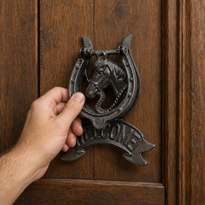 May include: A dark grey cast iron door knocker shaped like a horseshoe with a horse head in the centre and a banner at the bottom featuring the word WELCOME in raised capital letters, mounted on a vertical brown wooden door panel surface.