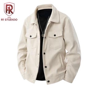Peut inclure: Une veste en velours côtelé beige avec un col pointu, des manches longues et deux poches poitrine à boutons noirs. La veste est portée sur une chemise noire et comprend un collier à chaîne argentée. Le logo R1 STUDIOO est visible en haut à gauche.