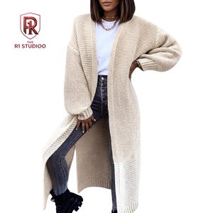 May include: A long, beige, open-front knit cardigan with long sleeves and a ribbed texture. The garment is worn over a plain white t-shirt and dark grey denim jeans. The logo for The R1 Studioo is visible in the upper left corner of the image.