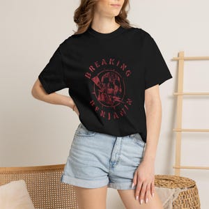 Könnte beinhalten: Ein schwarzes Kurzarm-T-Shirt mit einem roten Grafikdruck auf der Brust. Das Design zeigt einen Totenkopf in einem kreisförmigen Rahmen, begleitet von einem Blitzmotiv und dem Schriftzug "BREAKING BENJAMIN", der um das Motiv gebogen ist.