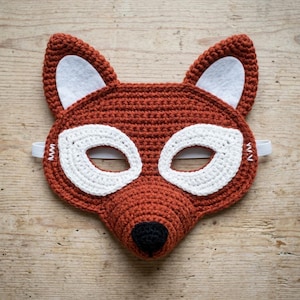 May include: A crocheted fox mask featuring a rust-orange yarn base with white felt inner ear accents and white crocheted eye patches. The mask includes a small black crocheted nose at the center and a white elastic band attached to the sides.