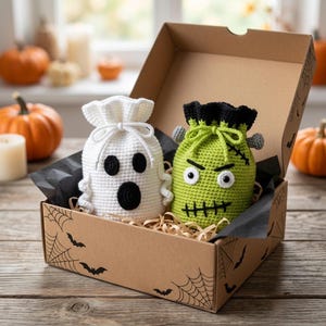 May include: A brown cardboard gift box decorated with black spiderwebs and bats contains two crocheted drawstring pouches. One pouch is white with a ghost face, and the other is lime green with a monster face, black hair, and grey bolts.