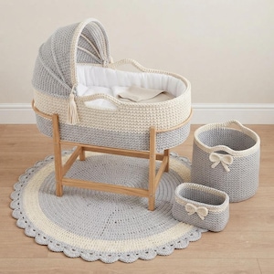 May include: A crochet bassinet with a grey and cream color scheme, featuring a hood and a wooden stand. The set includes a matching round scalloped rug, a large round storage basket, and a small rectangular basket, both adorned with cream bows.